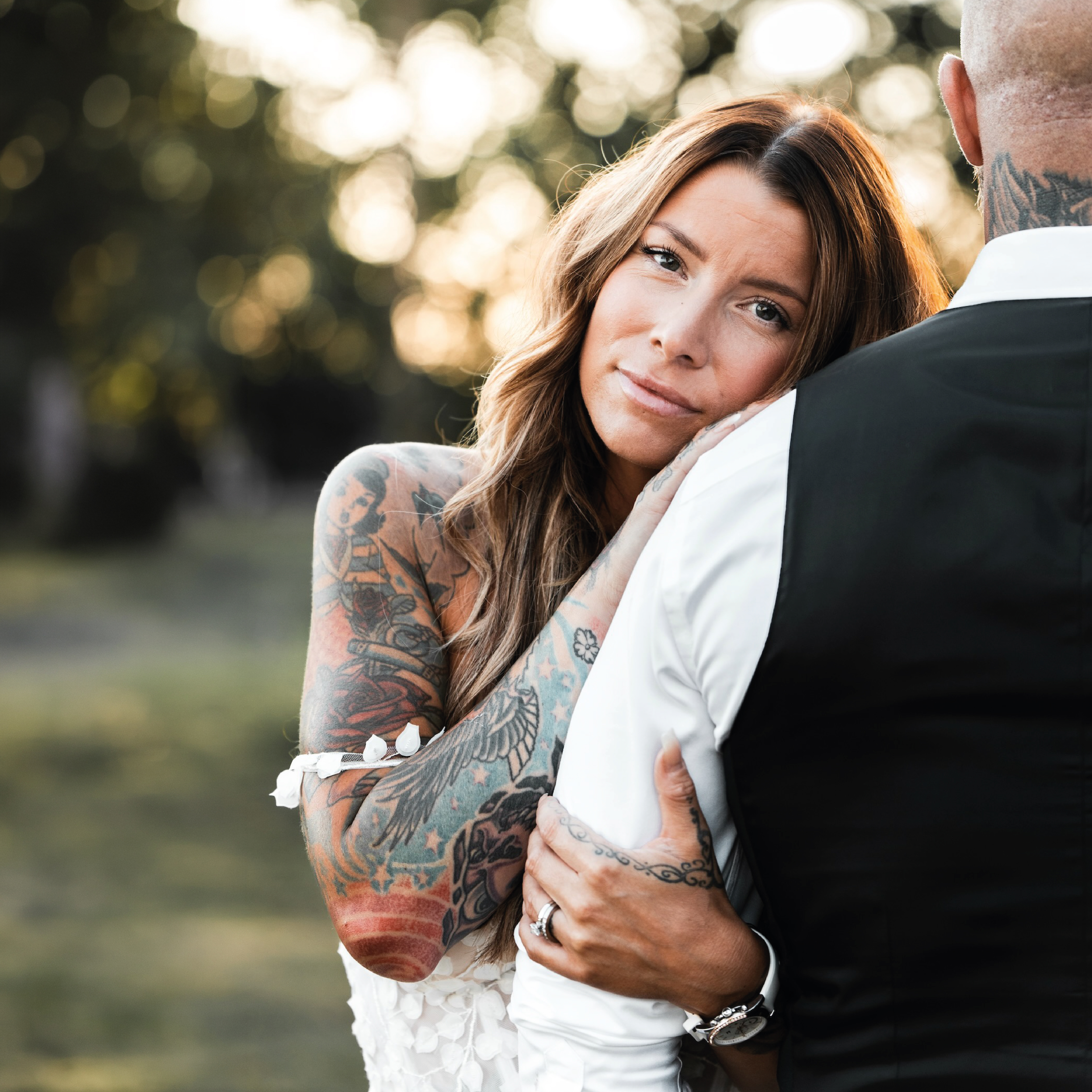En kvinna med långt ljusbrunt hår och synliga tatueringar på armarna lutar sitt huvud mot axeln på en man sedd bakifrån. Mannen bär en vit skjorta och en svart väst. Kvinnan blickar lugnt mot kameran och håller sin tatuerade hand på mannens rygg. Bakgrunden består av en mjukt suddig natur med ett varmt motljus (bokeh-effekt).